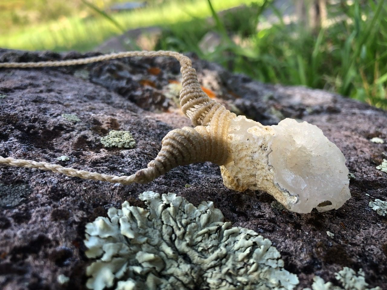 A small white geode and white knotwork string necklace rests on a lichen covered Boulder