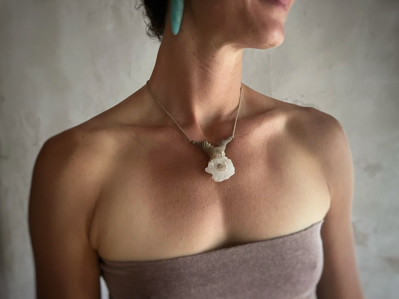 A woman wearing a pale purple top and a white thread and crystal necklace stands in front of a white mud wall