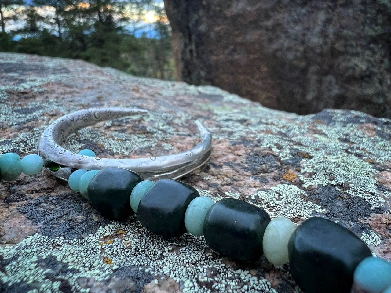 A necklace with a silver moon at the center and deep green jade and soft blue Amazonite beads rests on a lichen covered Boulder in the forest