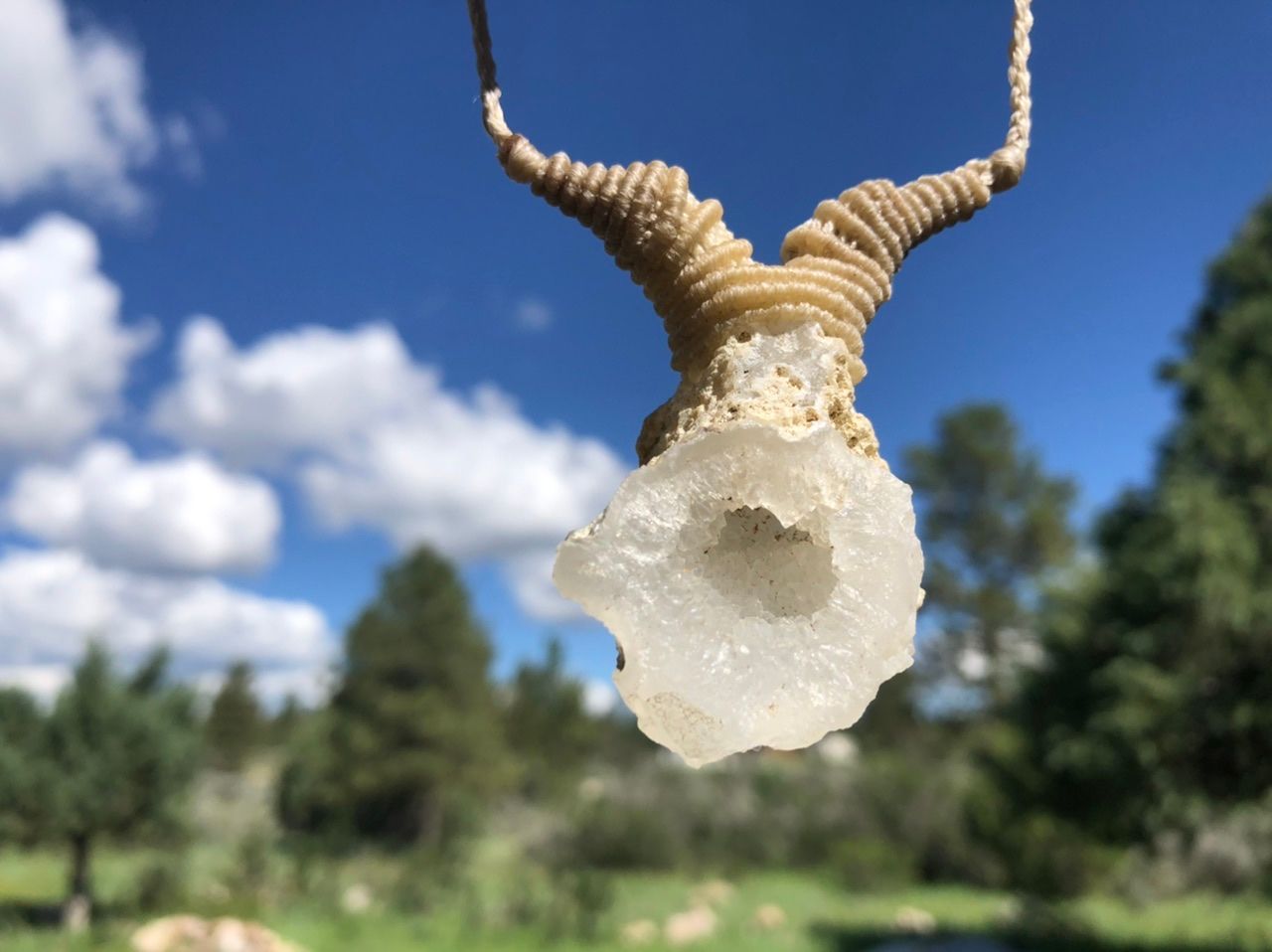  a white thread and crystal necklace Hangs in front of a blue sky and pine trees