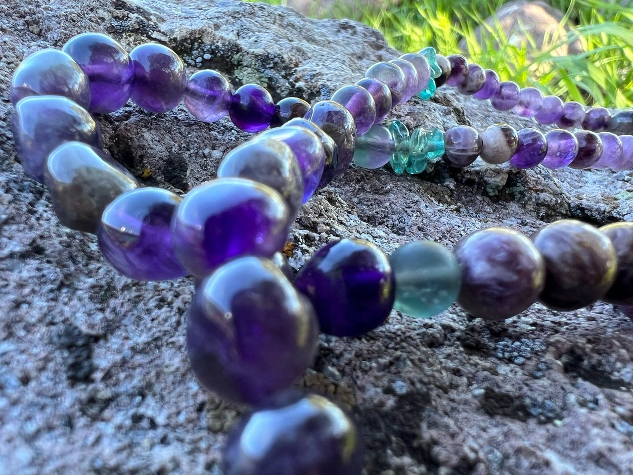 A necklace of purple, green and grey stones and crystals rests on a lichen covered Boulder 