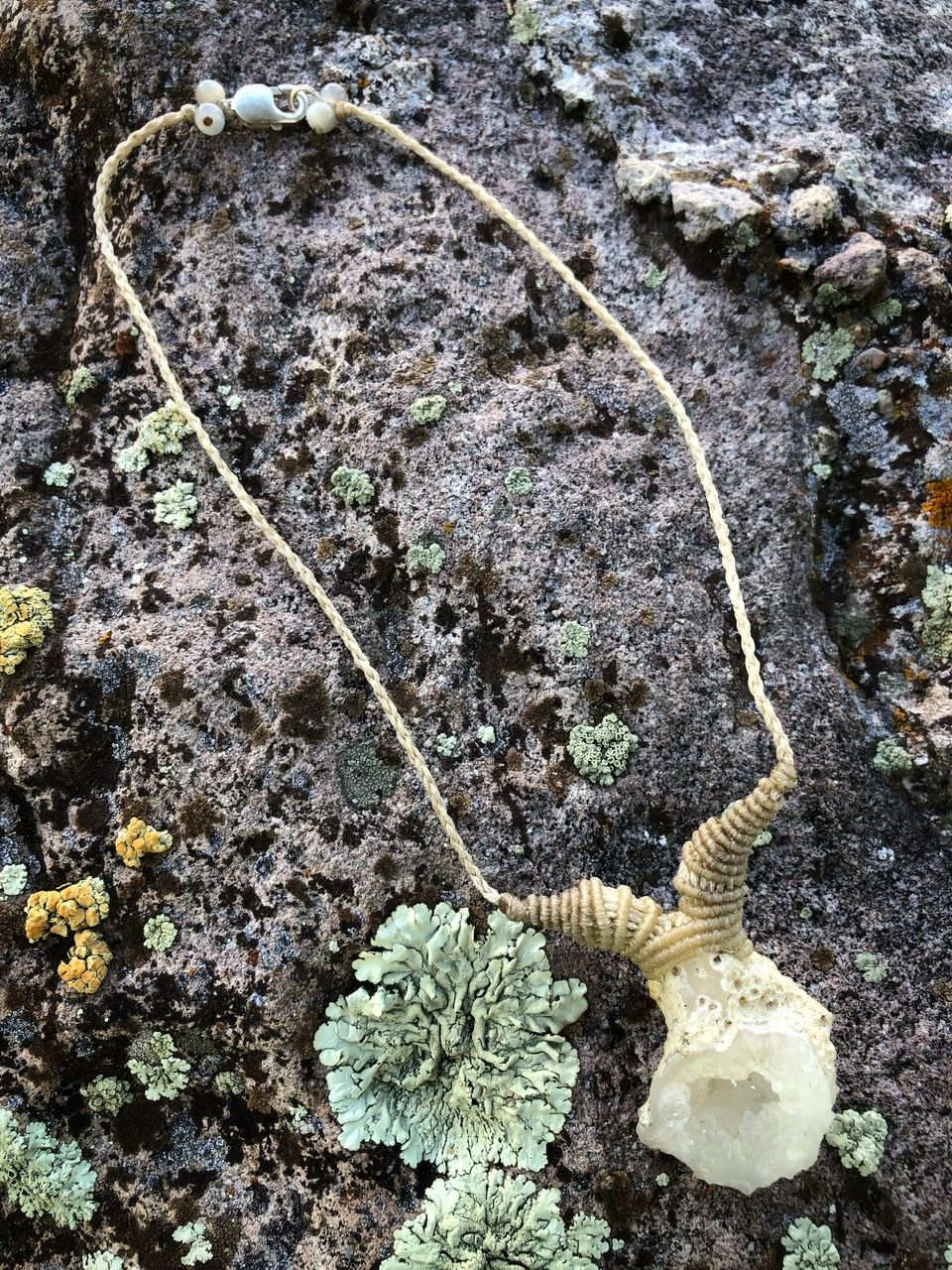 A small white geode and white knotwork string necklace rests on a lichen covered Boulder