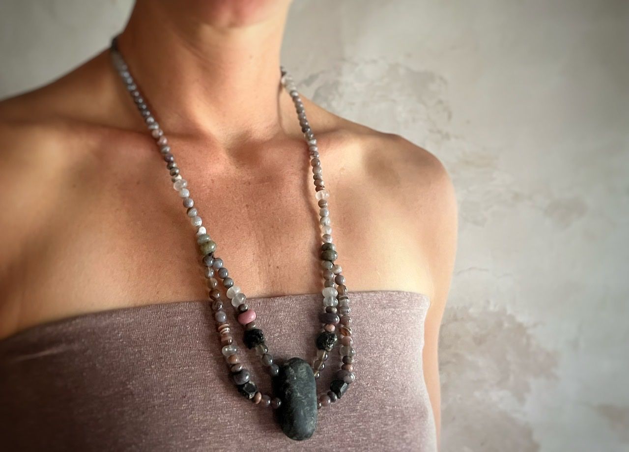 A woman wearing a pale purple top and a long stone necklace stands in front of a white mud wall