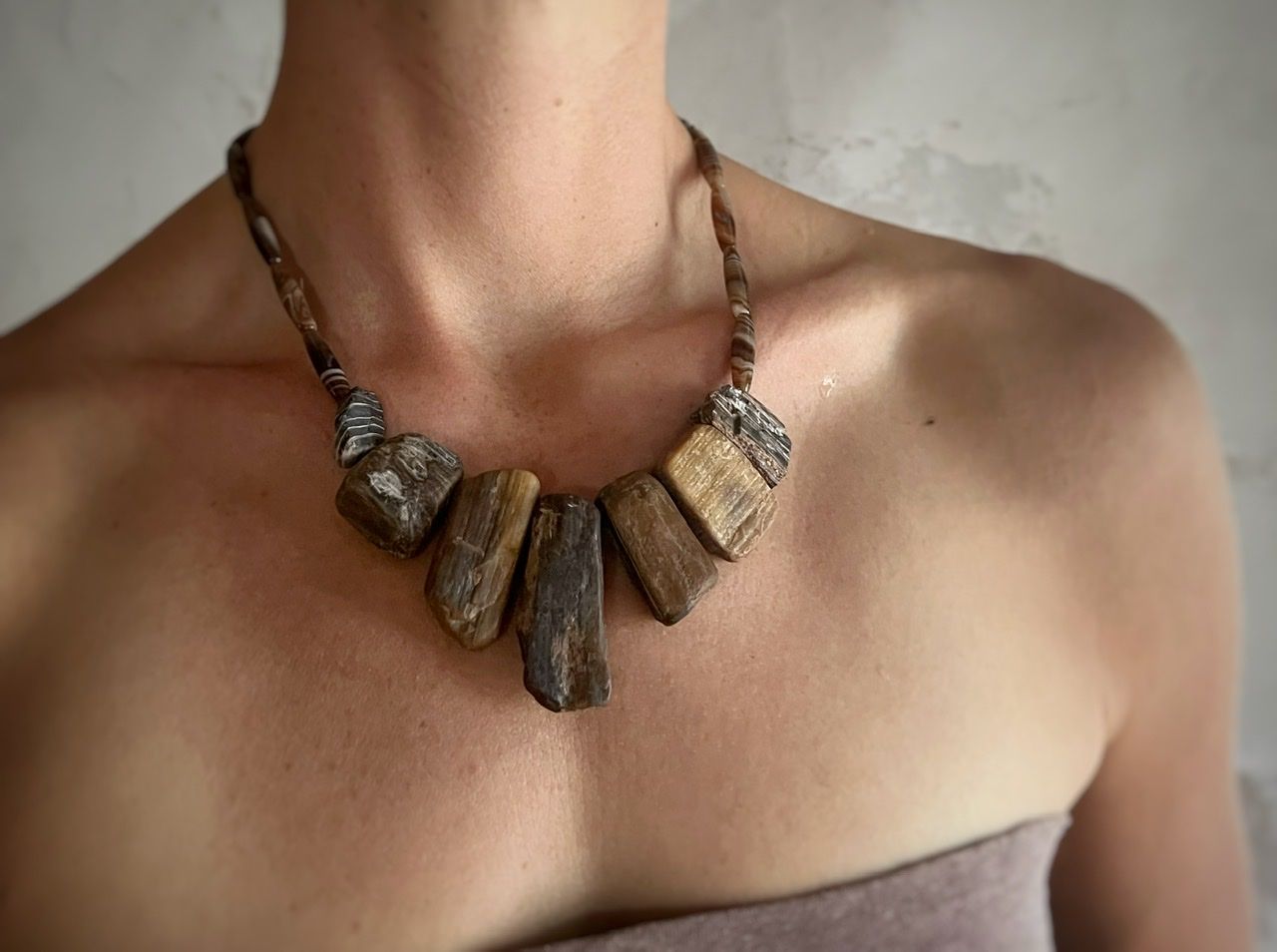 A woman wearing a pale purple top and a chunky petrified wood necklace stands in front of a white mud wall