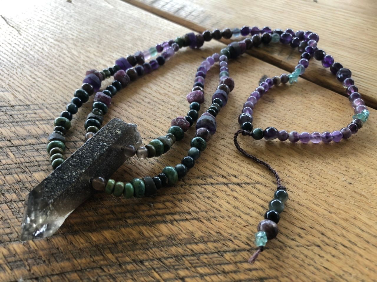A necklace of purple, green and grey stones and crystals rests on an oak table