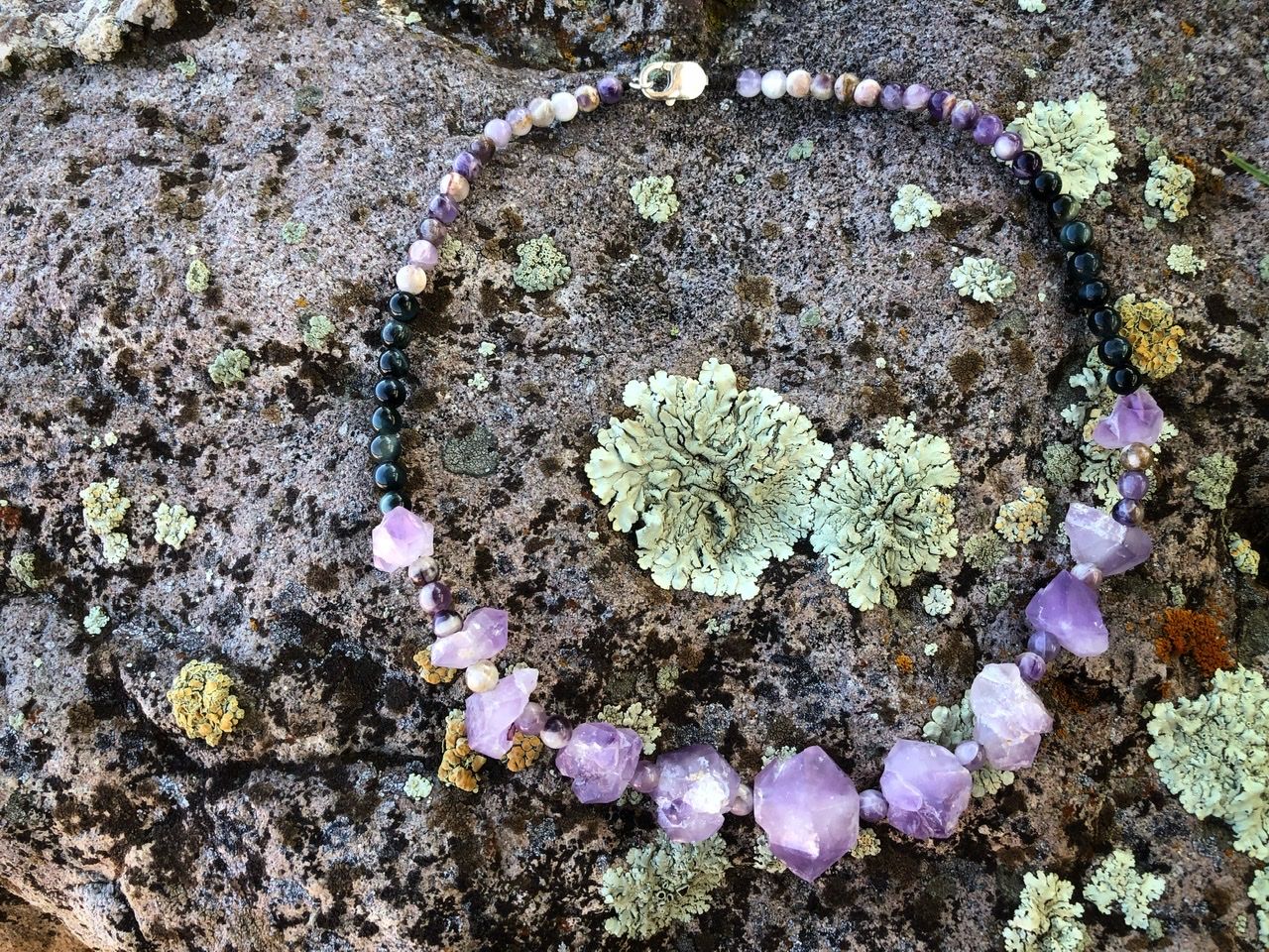 A purple amethyst crystal and black cats eye necklace rests on a lichen covered Boulder