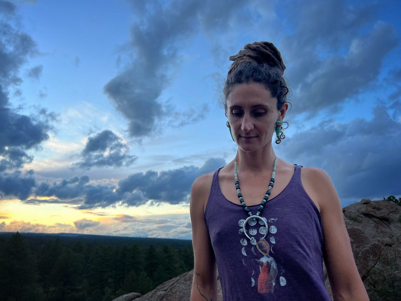 A woman wears A necklace with a silver moon at the center and deep green jade and soft blue Amazonite beads on a mountaintop at sunset