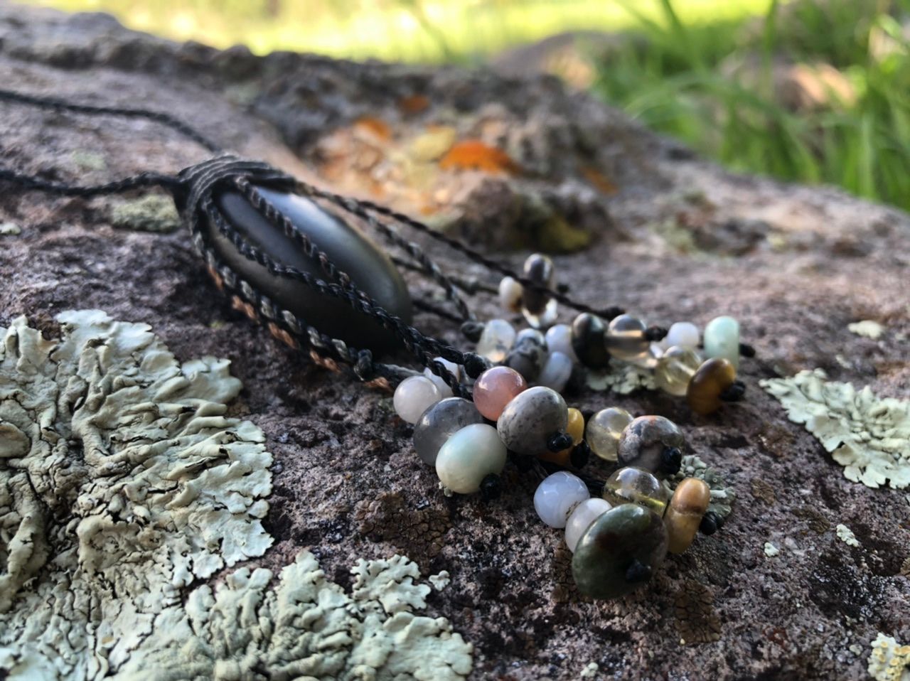 A smooth black ocean tumbled stone talisman necklace with braided dangling beads, rests on a lichen covered Boulder