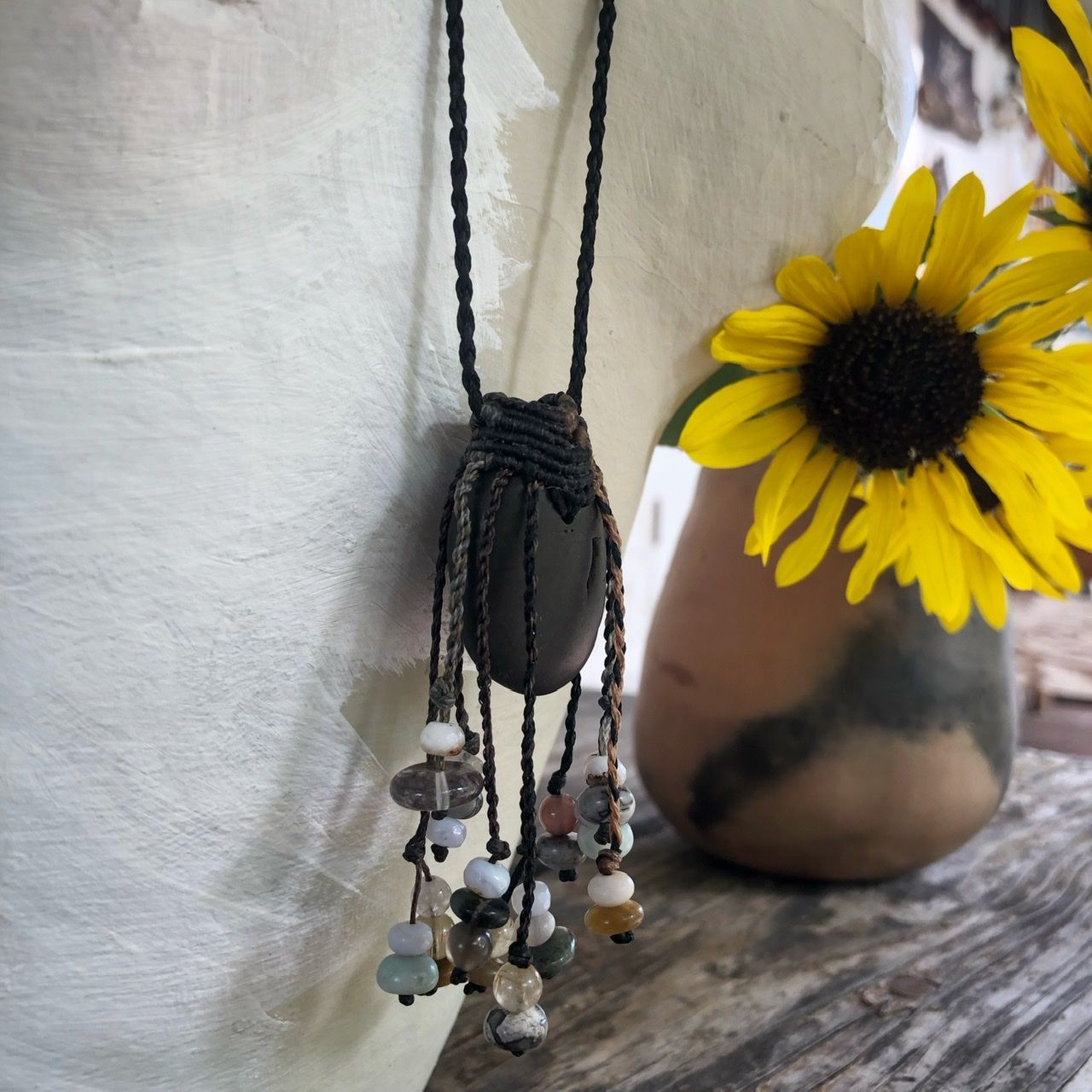 A smooth black ocean tumbled stone talisman necklace with braided dangling beads, rests on a white mannequin on a wood table with sunflowers in the background 