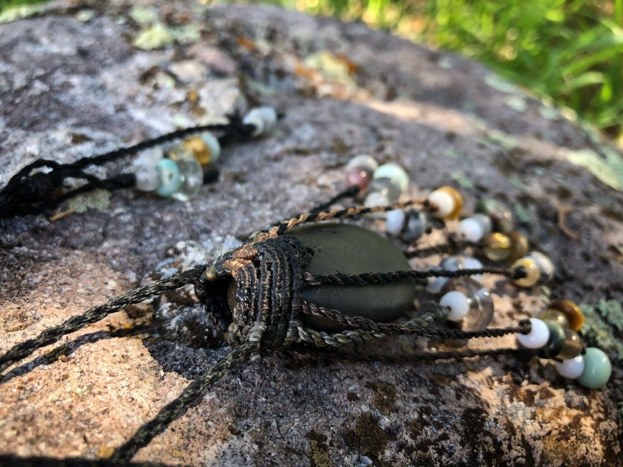 A smooth black ocean tumbled stone talisman necklace with braided dangling beads, rests on a lichen covered Boulder