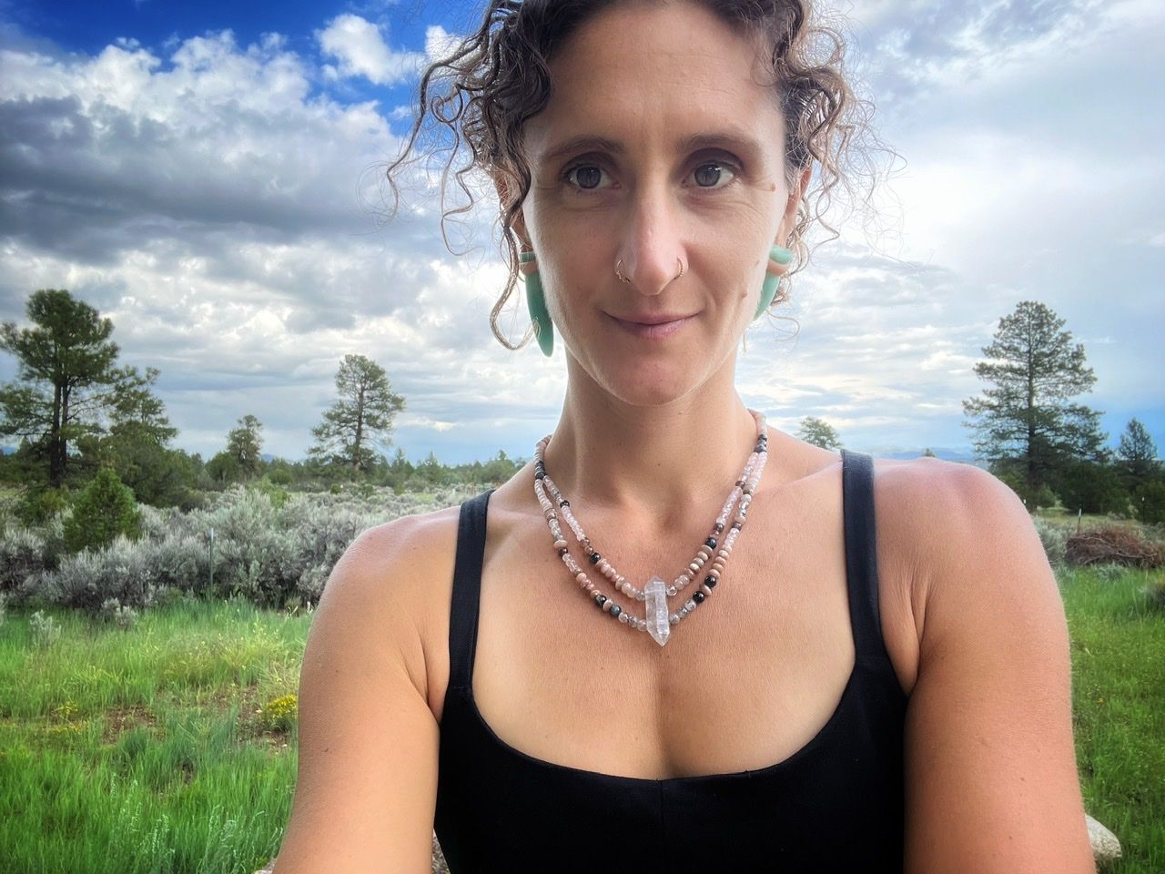 A woman wearing a stone beaded necklace in pale pinks, dusty pinks, black and grays standing in a green field with ponderosa pines behind 
