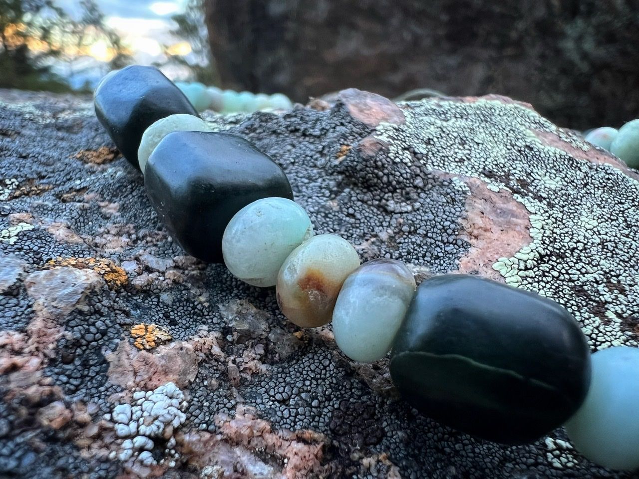 A necklace with a silver moon at the center and deep green jade and soft blue Amazonite beads rests on a lichen covered Boulder in the forest