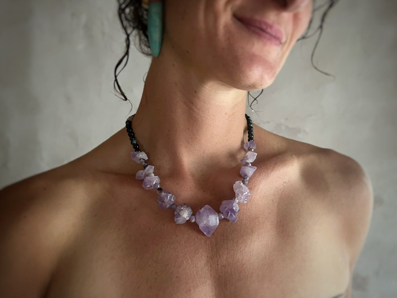 A woman wearing a pale purple top and a short amethyst crystal necklace stands in front of a white mud wall