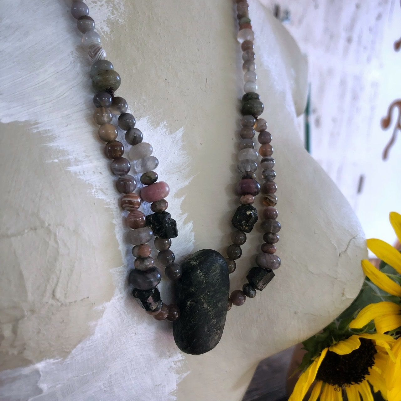 A two strand crystal and stone necklace rests on a white mannequin on a wood table with sunflowers in the background 