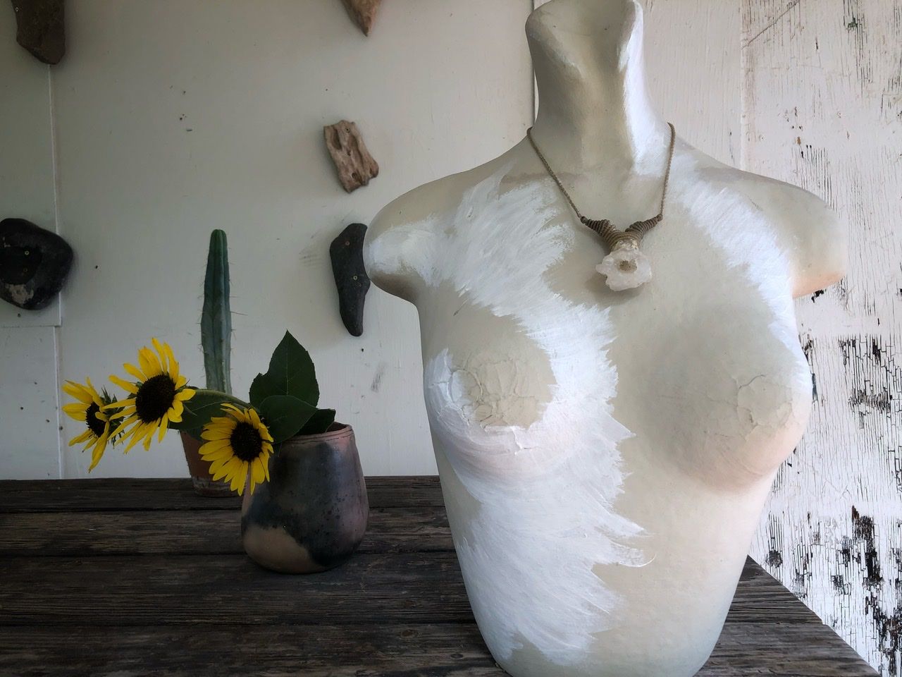 A white crystal geode and white string necklace rests on a white mannequin on a wood table with sunflowers in the background 