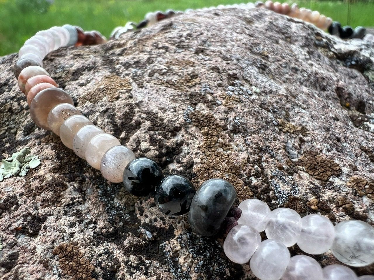 a stone beaded necklace in pale pinks, dusty pinks, black and grays lays on a lichen covered stone