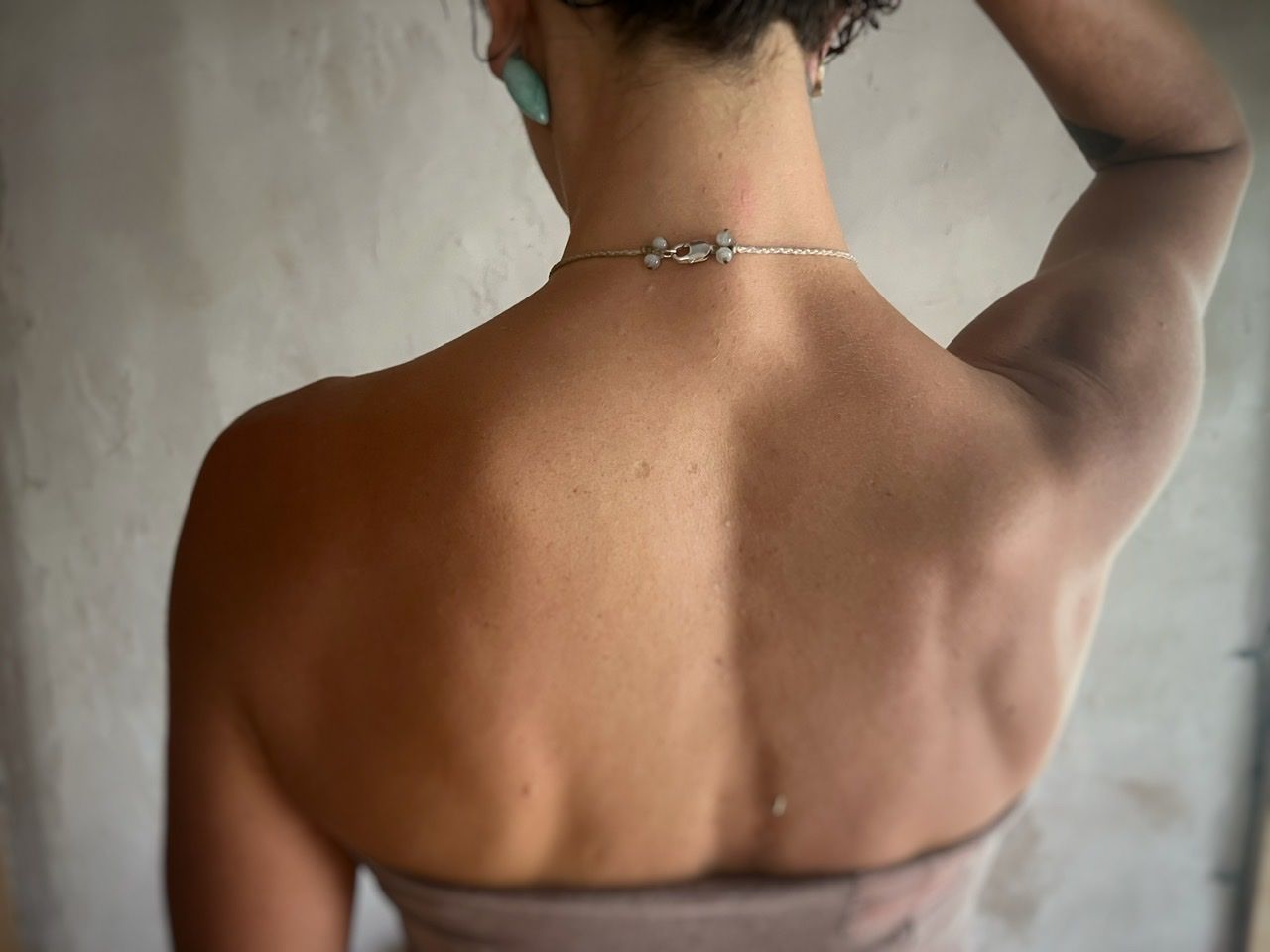 A woman wearing a pale purple top and a white thread and crystal necklace stands in front of a white mud wall