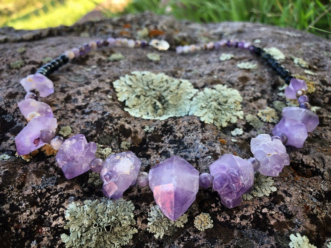 A purple amethyst crystal and black cats eye necklace rests on a lichen covered Boulder