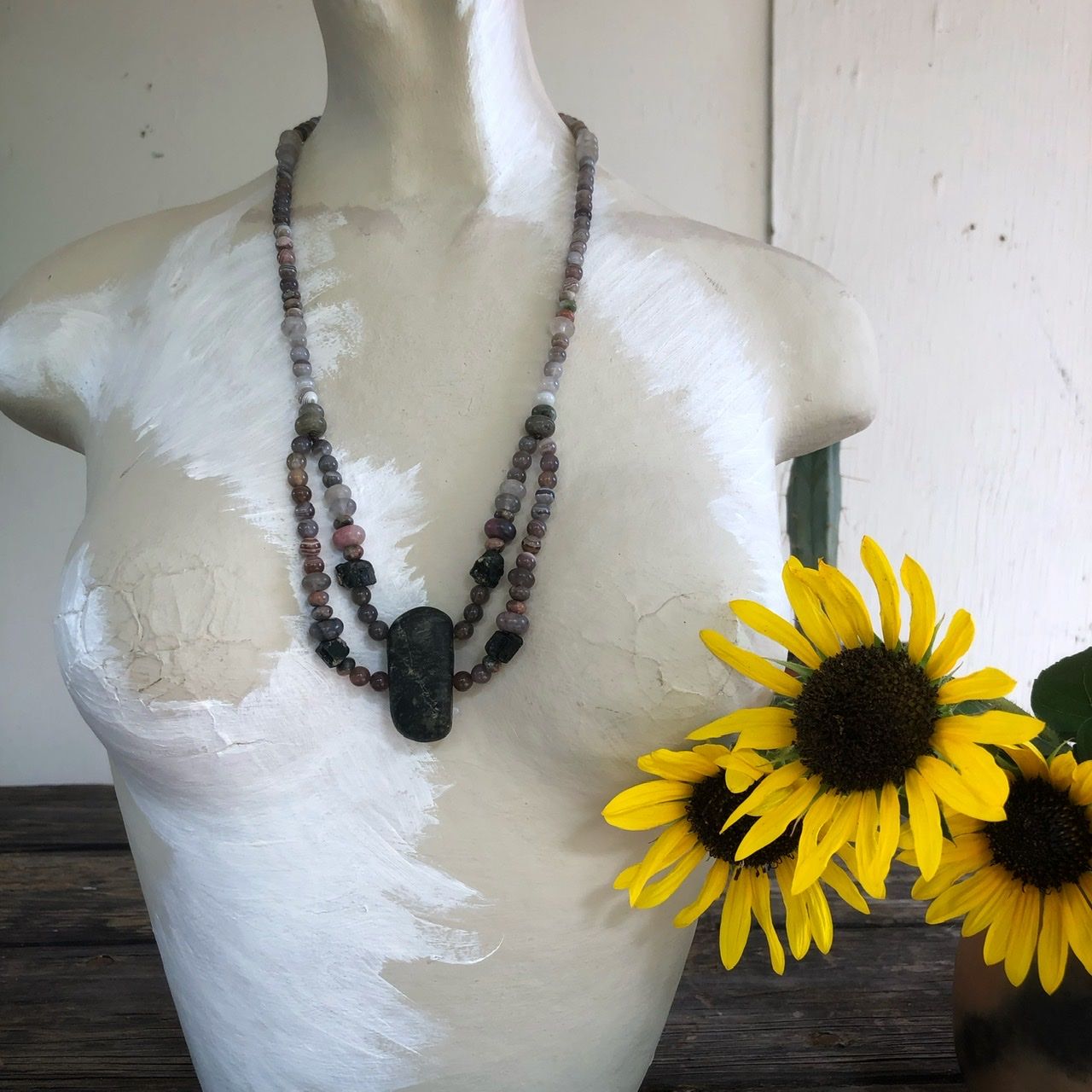 A two strand crystal and stone necklace rests on a white mannequin on a wood table with sunflowers in the background 