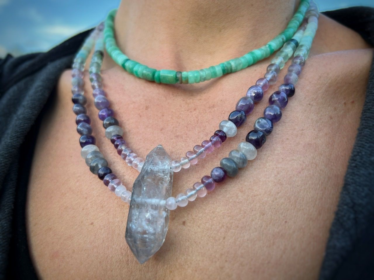 A detail of a woman wearing A necklace of clear, pink and grey Quartz, purple amethyst, green fluorite and blue chalcedony