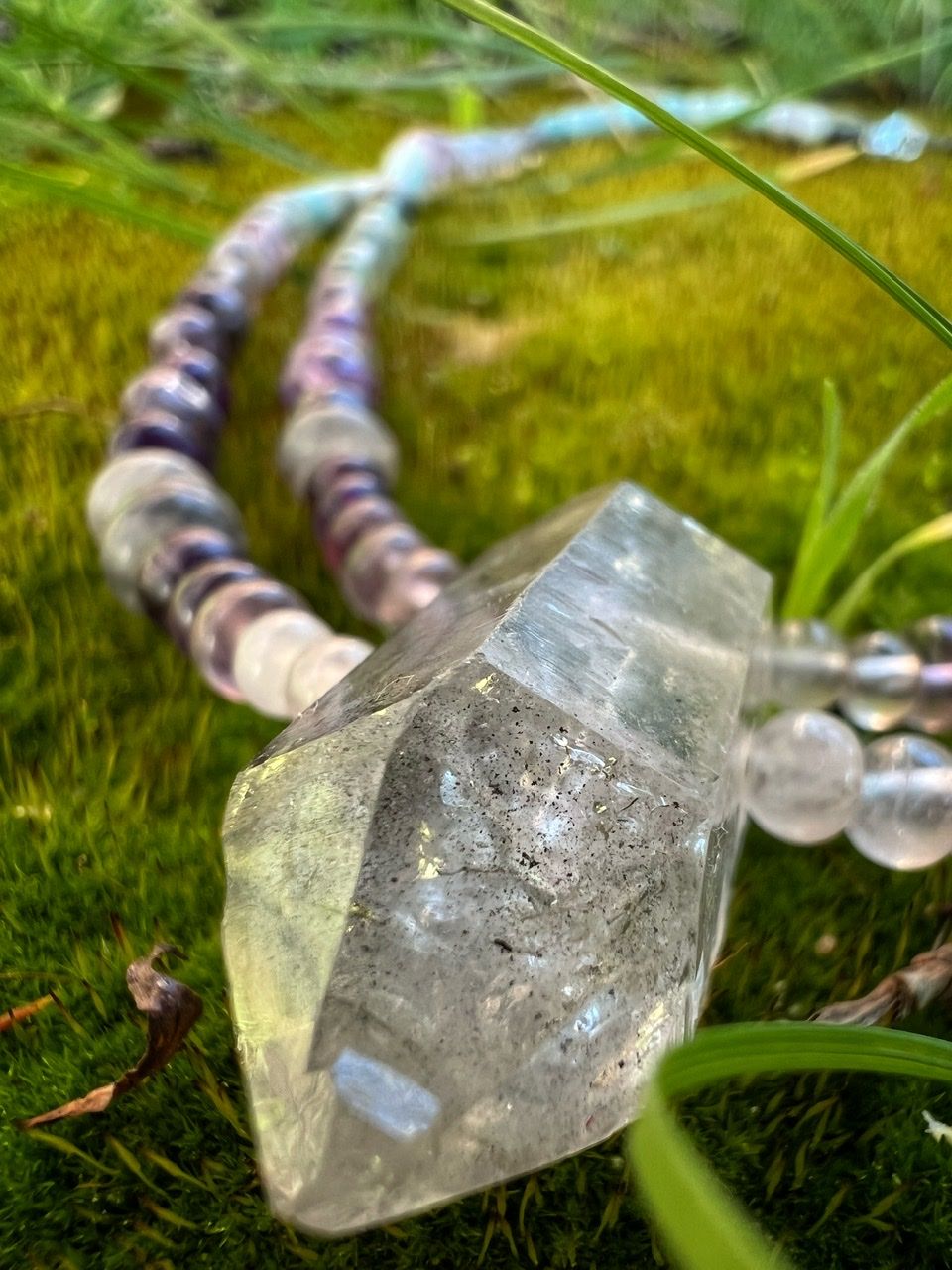 A necklace of clear, pink and grey Quartz, purple amethyst, green fluorite and blue chalcedony rests on lush green moss