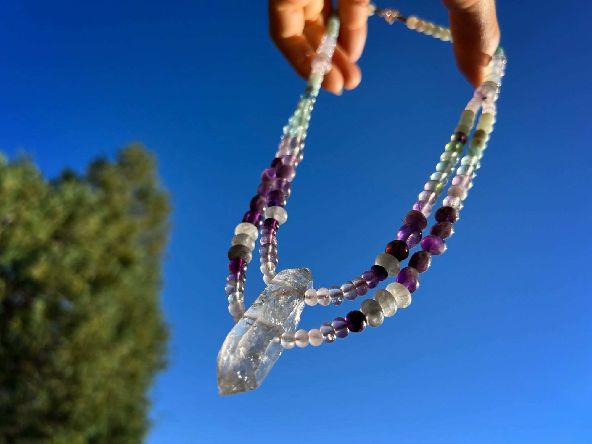 A necklace of clear, pink and grey Quartz, purple amethyst, green fluorite and blue chalcedony hangs against a blue sky 