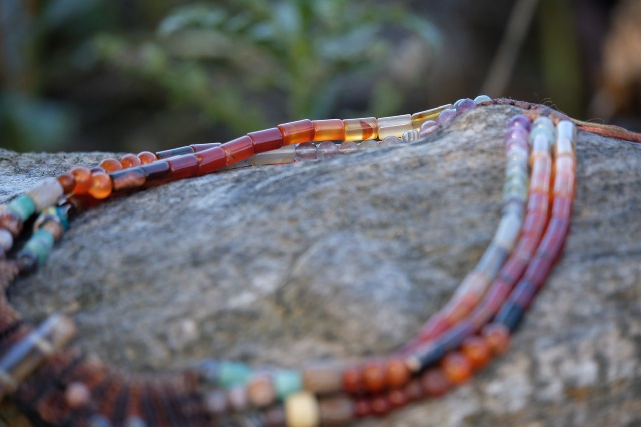 A handmade necklace of red, green, orange, purple precious stones and knotwork lay on a gray log