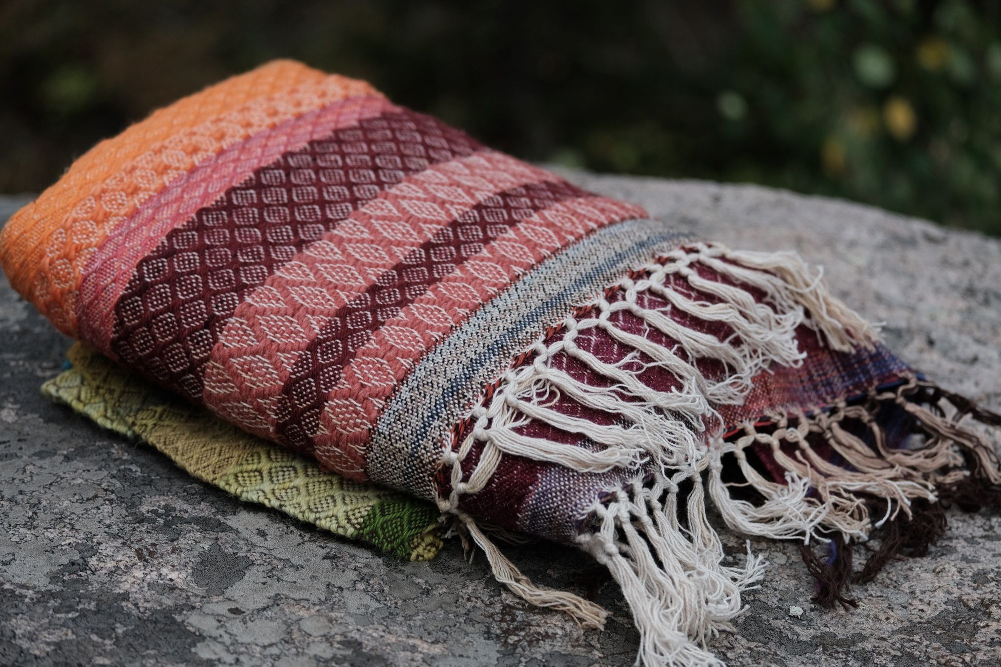 A handwoven rainbow striped blanket lays folded on a lichen covered rock in the forest
