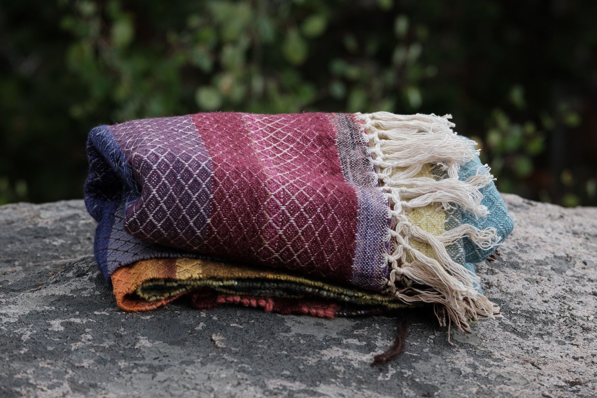 A handwoven rainbow striped blanket lays folded on a lichen covered rock in the forest