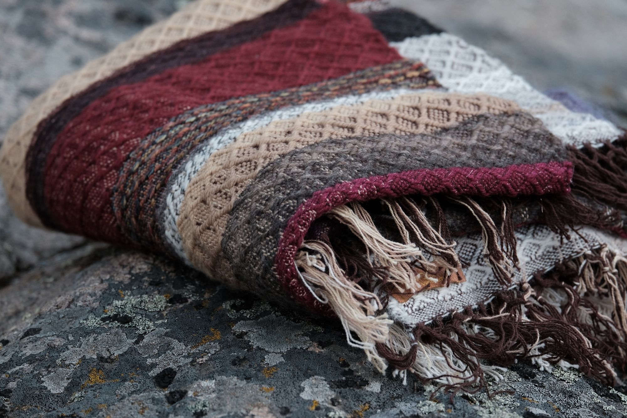 A maroon, black, purple, tan and white handwoven diamond pattern blanket lays folded on a lichen covered boulder