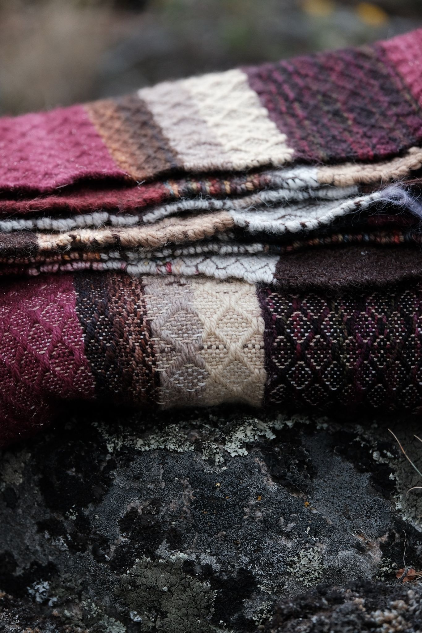 A maroon, black, purple, tan and white handwoven diamond pattern blanket lays folded on a lichen covered boulder