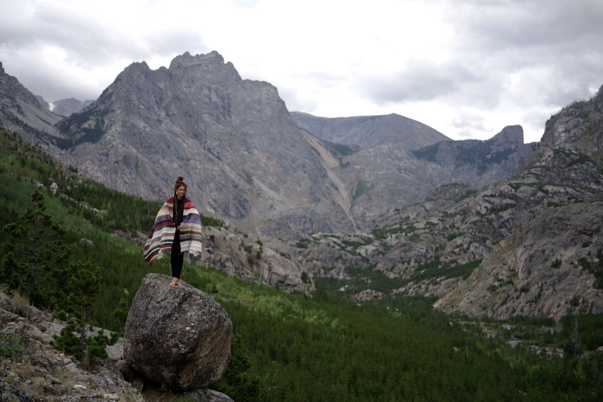 A woman stands atop a boulder wearing a handwoven striped blanket, in a majestic mountain valley