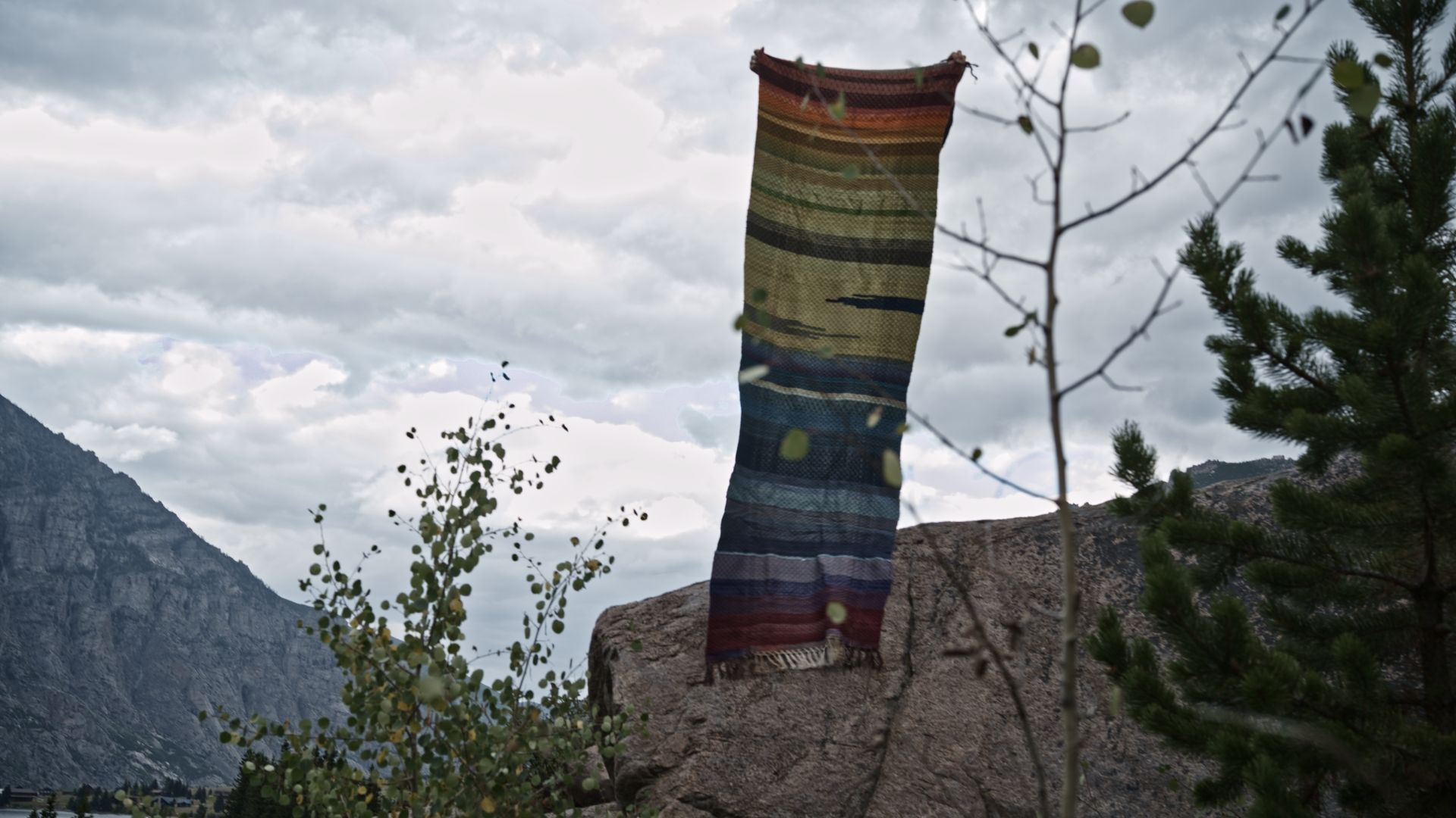 A woman stands atop a boulder wearing a handwoven rainbow striped blanket, in a majestic mountain valley