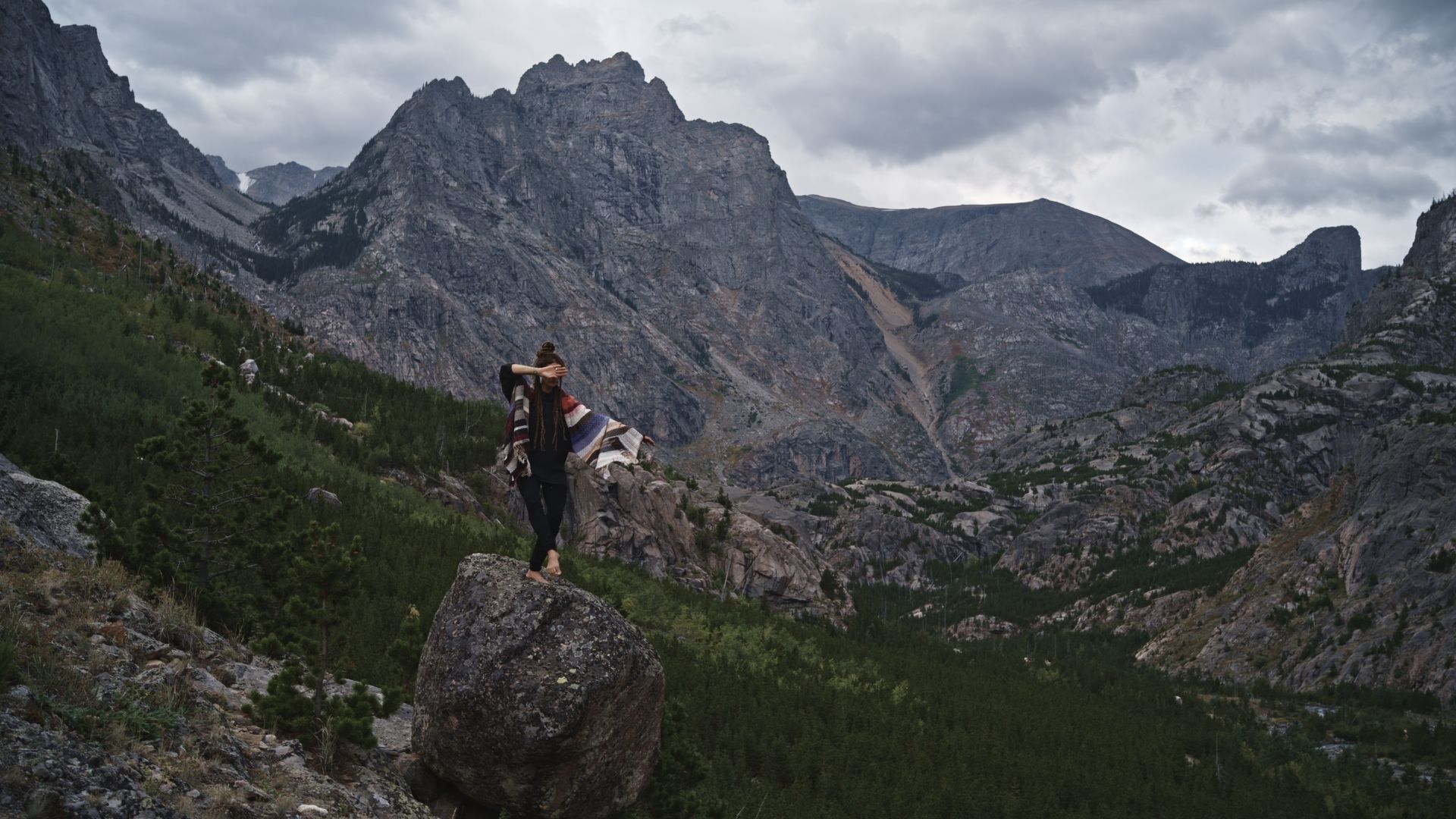 A woman stands atop a boulder wearing a handwoven striped blanket, in a majestic mountain valley