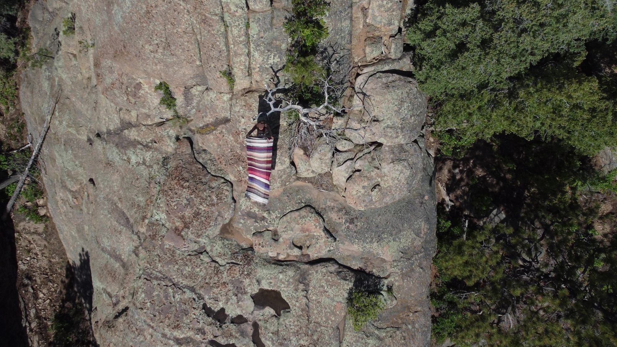 A view from above of a woman wearing a maroon, purple, blue and tan handwoven blanket, standing on a giant monolithic rock in the forest