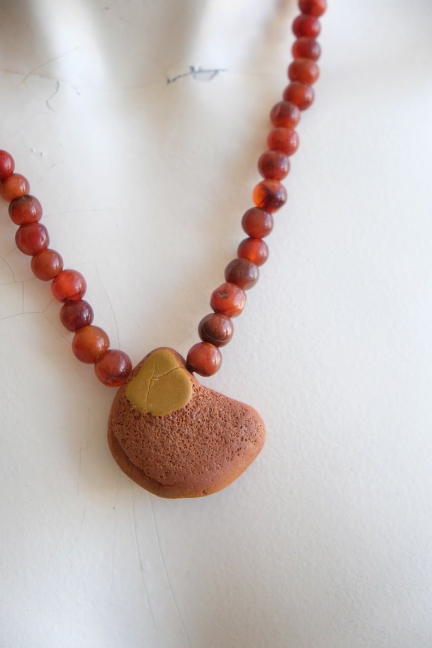 A white color female form mannequin wears a sculptural stone necklace in red, orange and yellow ochre colors 