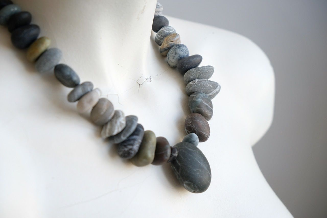 A white color female form mannequin wears a sculptural necklace of smooth dark blue grey stones