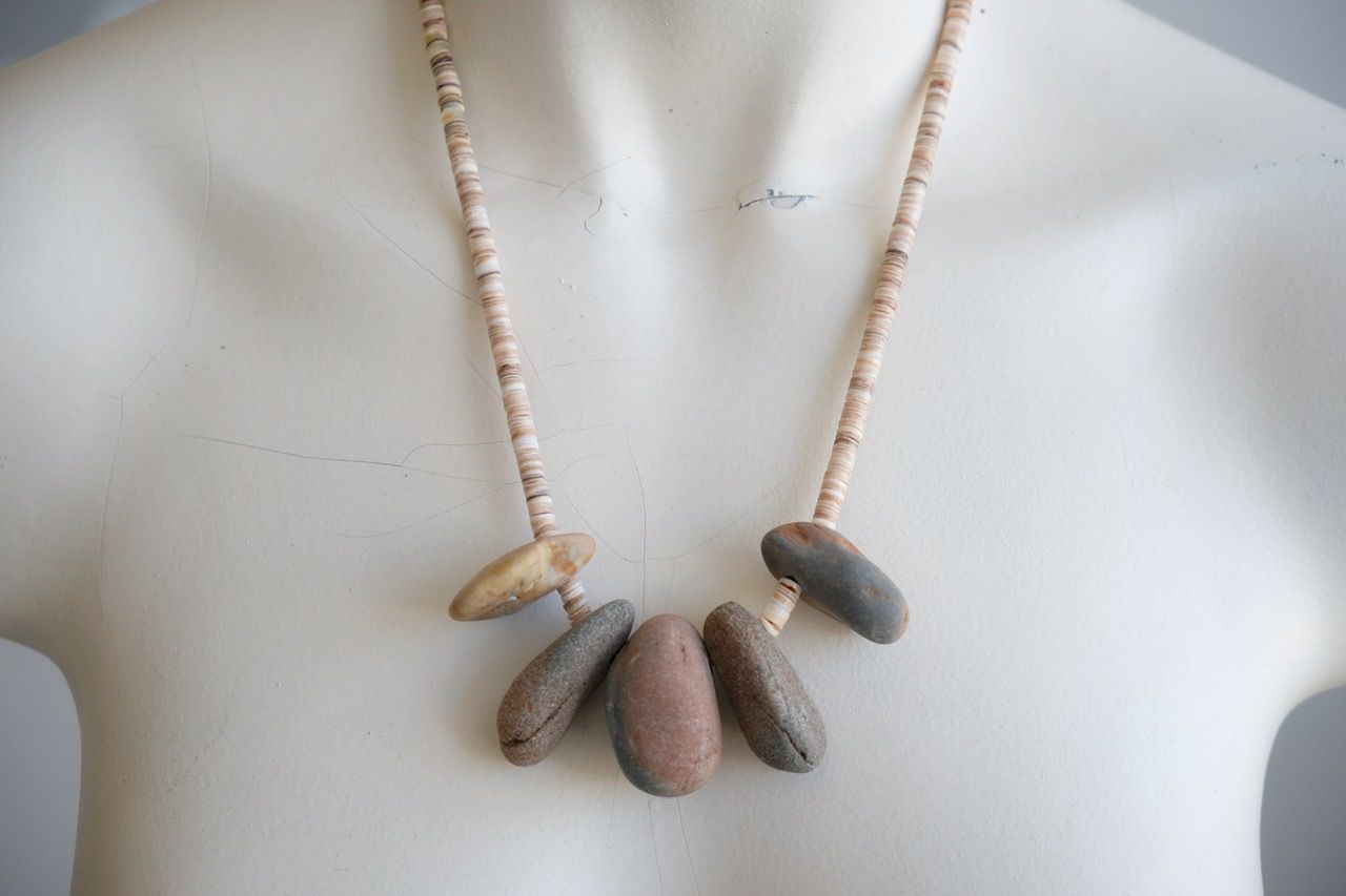 A white female form mannequin wears a sculptural necklace of ocean tumbled stones In greys, brown, red and white