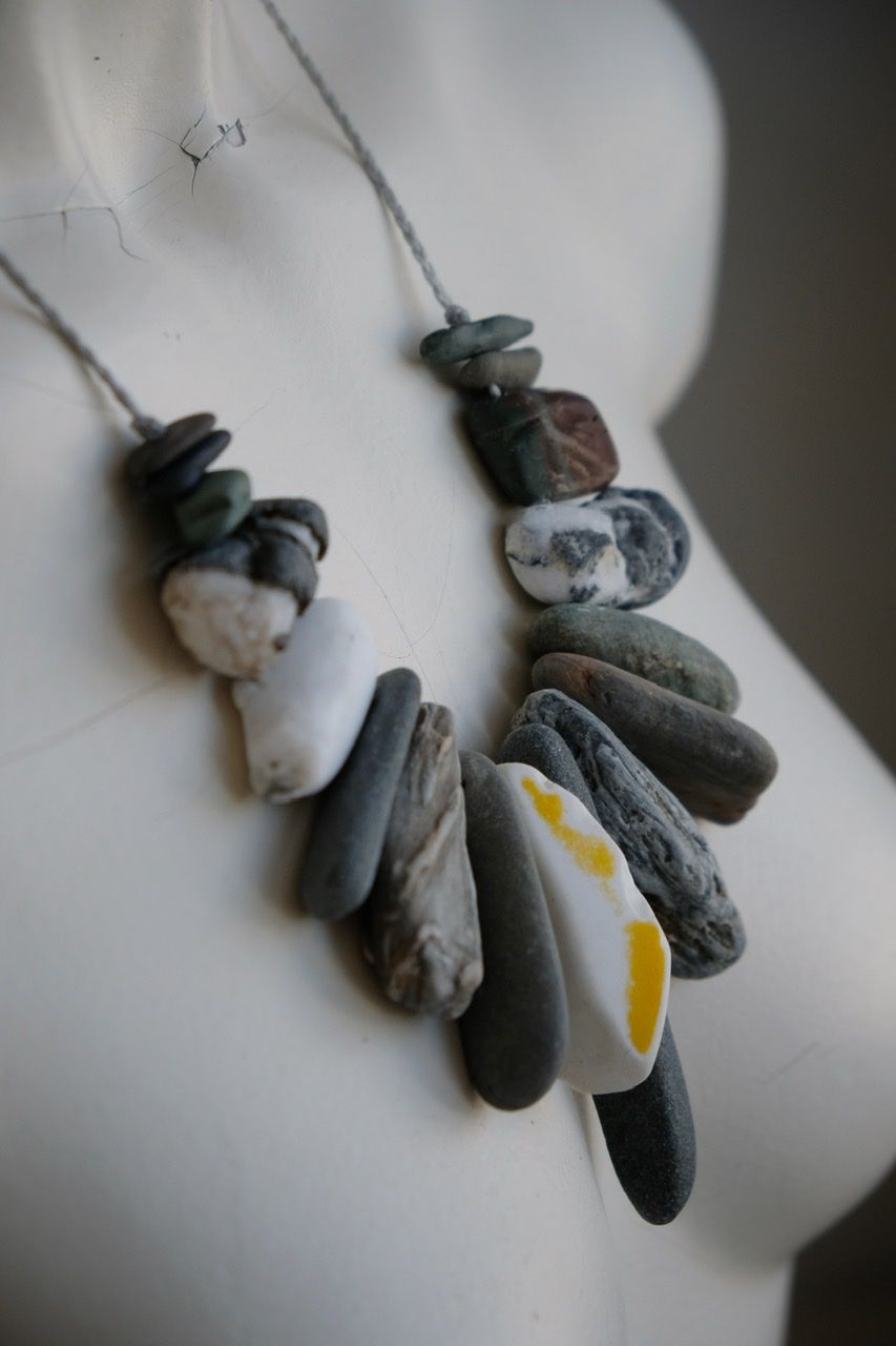 A white colored female form mannequin wears a sculptural necklace of gray braided twine with rough stones hanging from it.