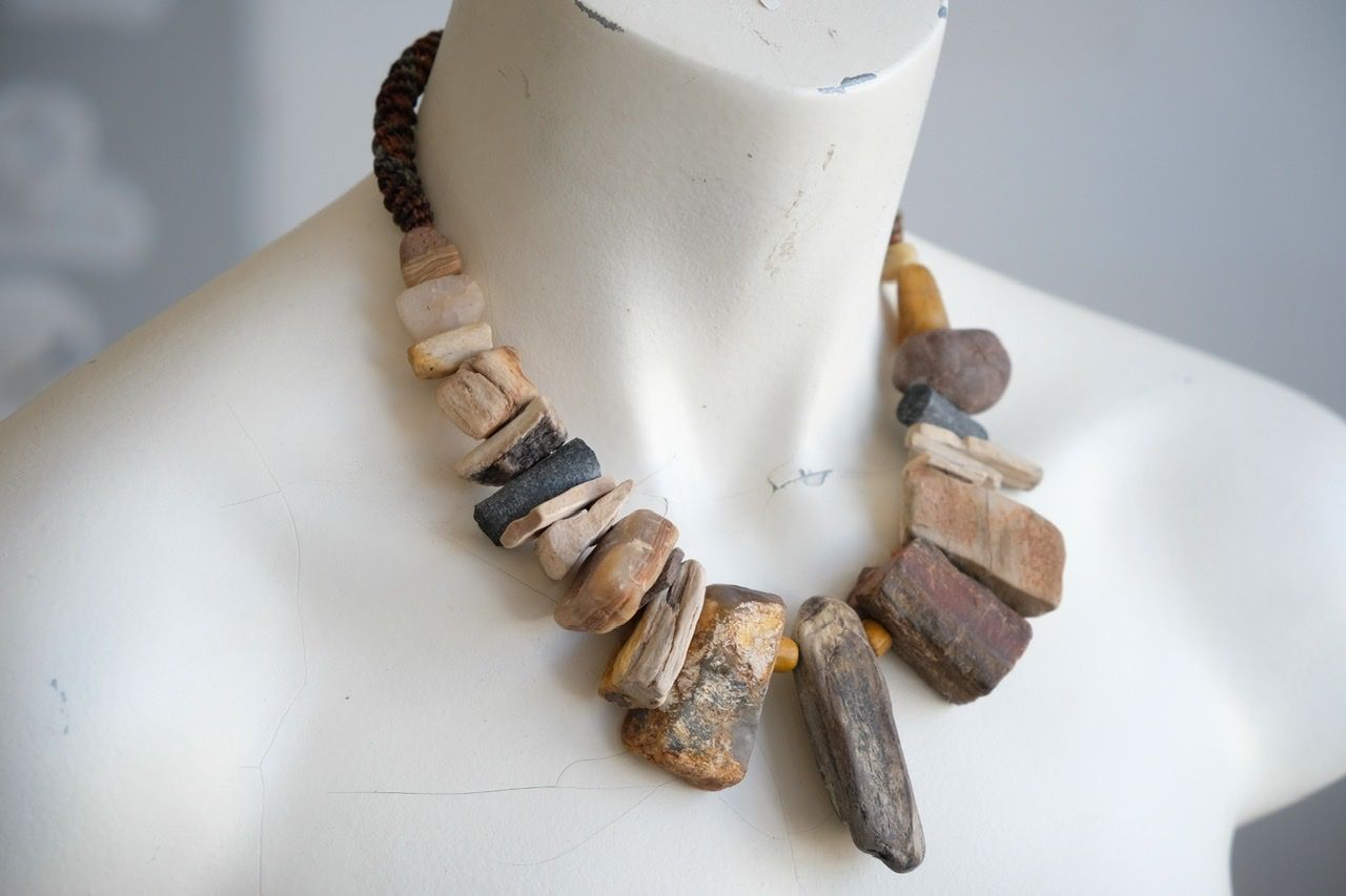 A white colored female form mannequin wears a sculptural necklace of petrified wood, smooth stones, yellow glass beads and a knot work tube