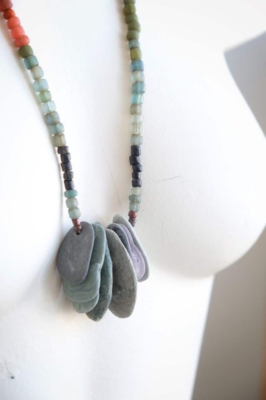 A white colored female form mannequin wears a sculptural necklace of thin grey stones and multicolored glass beads. 