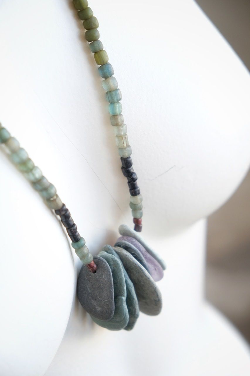 A white colored female form mannequin wears a sculptural necklace of thin grey stones and multicolored glass beads. 