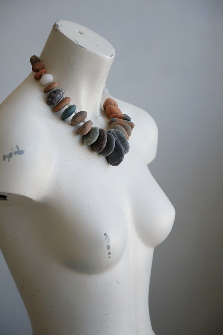 A white female form mannequin wears a sculptural necklace of ocean tumbled stones In greys, brown, red and white