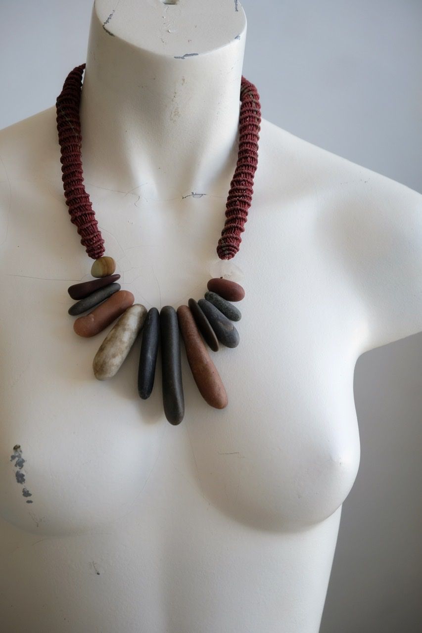A white colored female form mannequin wears a sculptural necklace of multicolored smooth stones and raspberry colored basketweaving