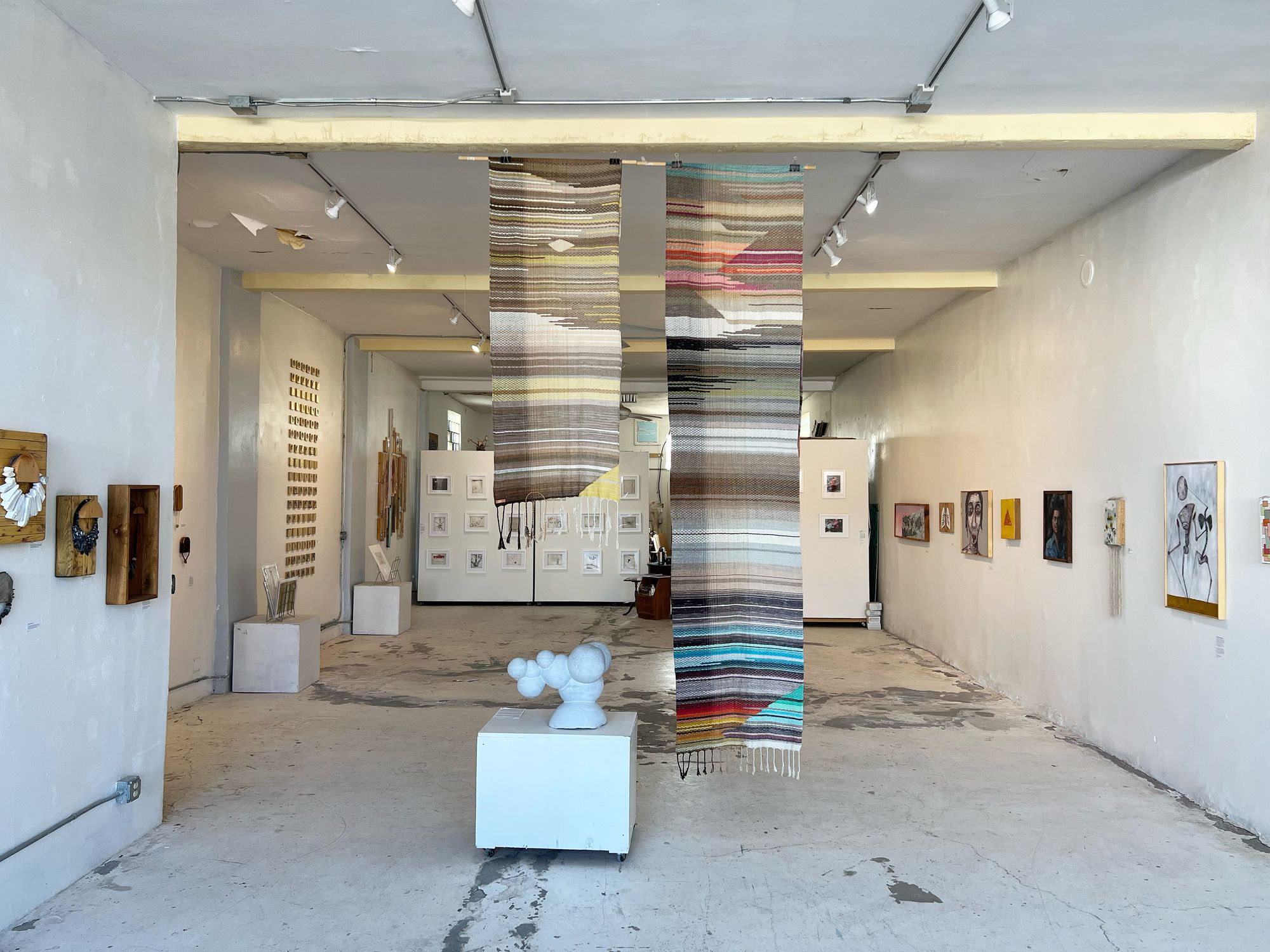 Handwoven natural and rainbow colored fabric hanging in the middle of a gallery with art on the white walls all around and a marble sculpture next to it