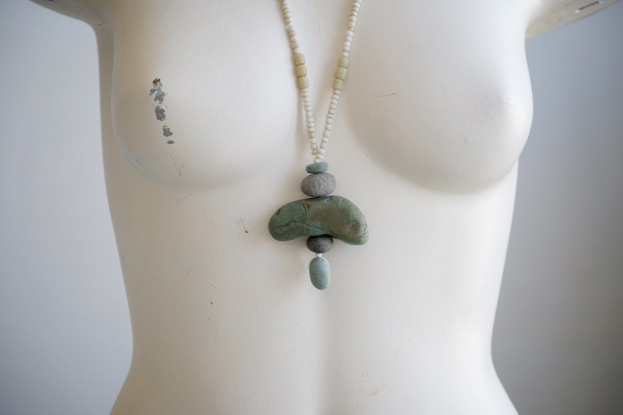 A white colored female form mannequin wears a sculptural necklace of blue green river tumbled stones and white glass beads