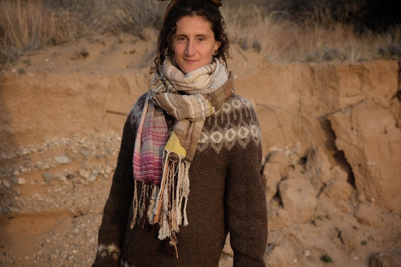 A woman in a brown and tan sweater stands in a sandy desert landscape wearing a shawl