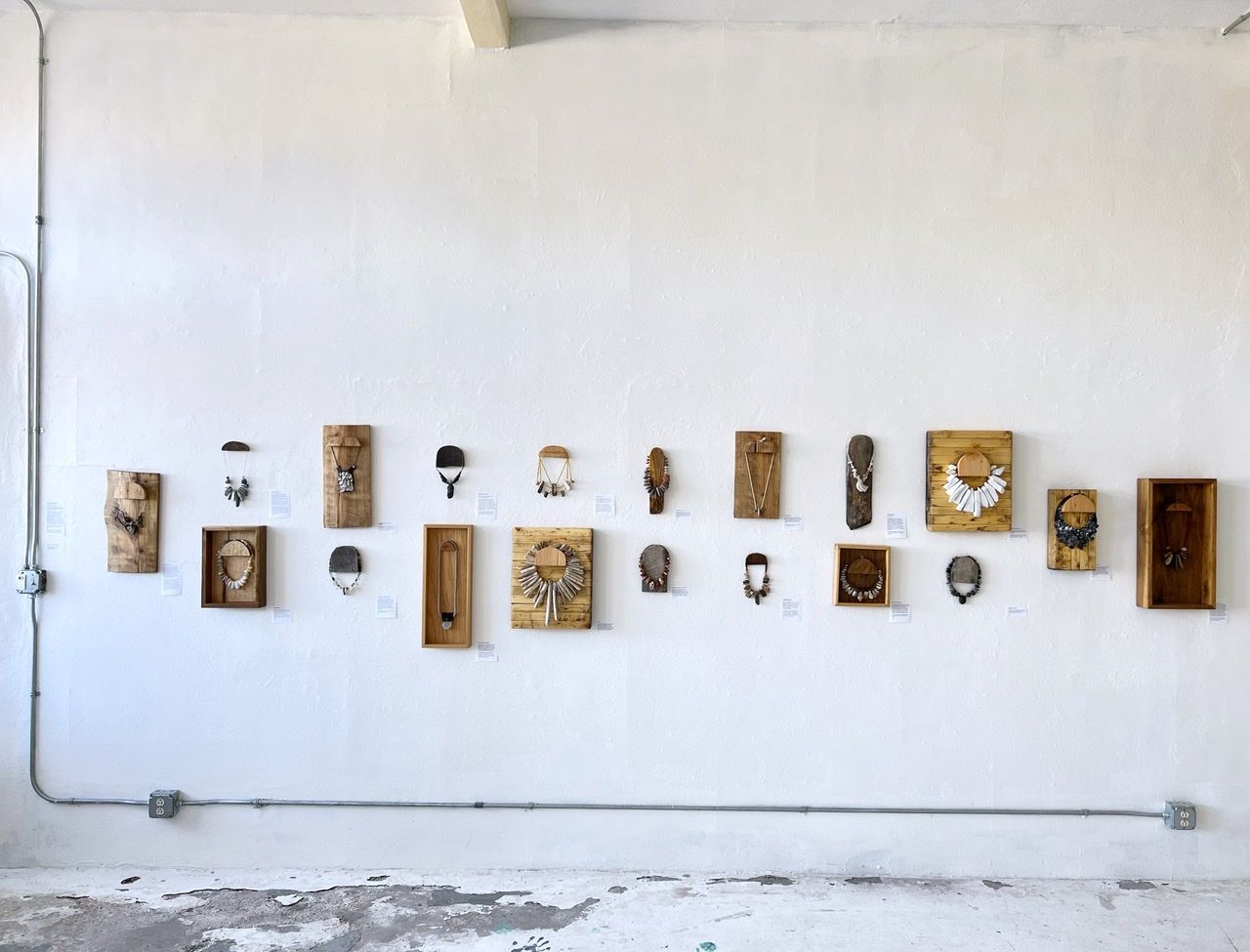 19 necklaces of stone, beads, string and wood hang on wooden frames on a white gallery wall
