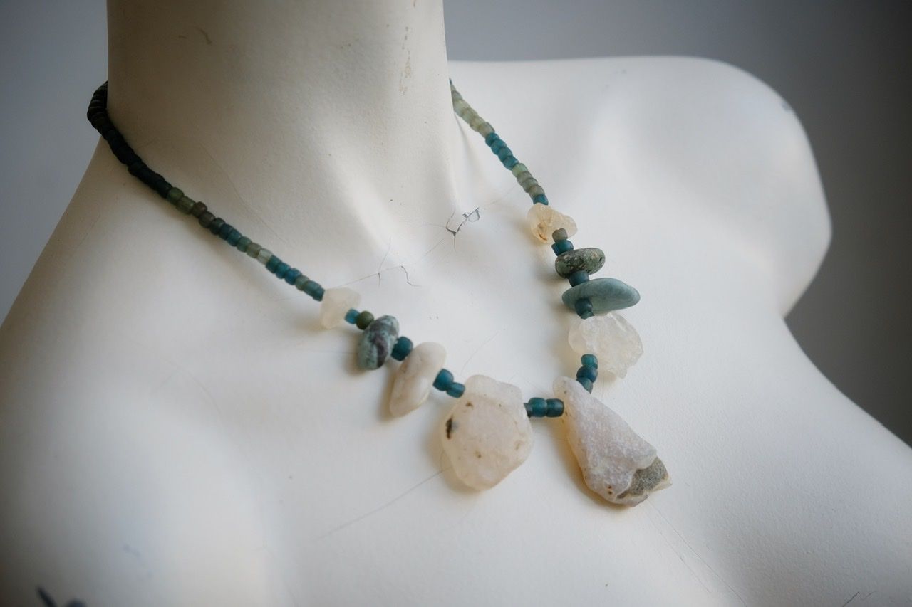 A white colored female form mannequin wears a sculptural necklace of white and blue river tumbled stones and turquoise glass beads