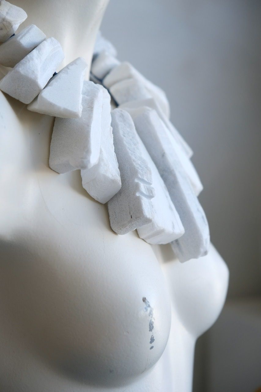 A white colored female form mannequin wears a sculptural necklace of large wedges of white marble and blue string